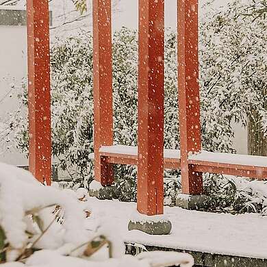 Ein Paar spaziert im Schnee unter einer roten Pergola im Chinesischen Garten, umgeben von verschneiten Bambussträuchern.