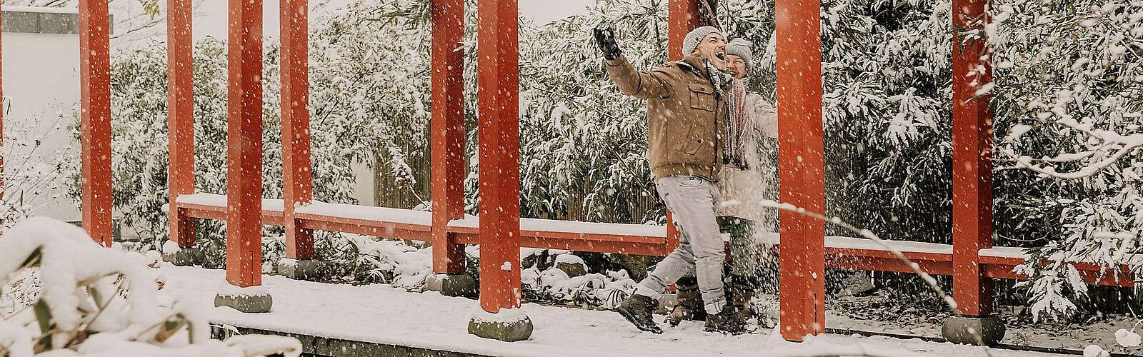 Ein Paar spaziert im Schnee unter einer roten Pergola im Chinesischen Garten, umgeben von verschneiten Bambussträuchern.